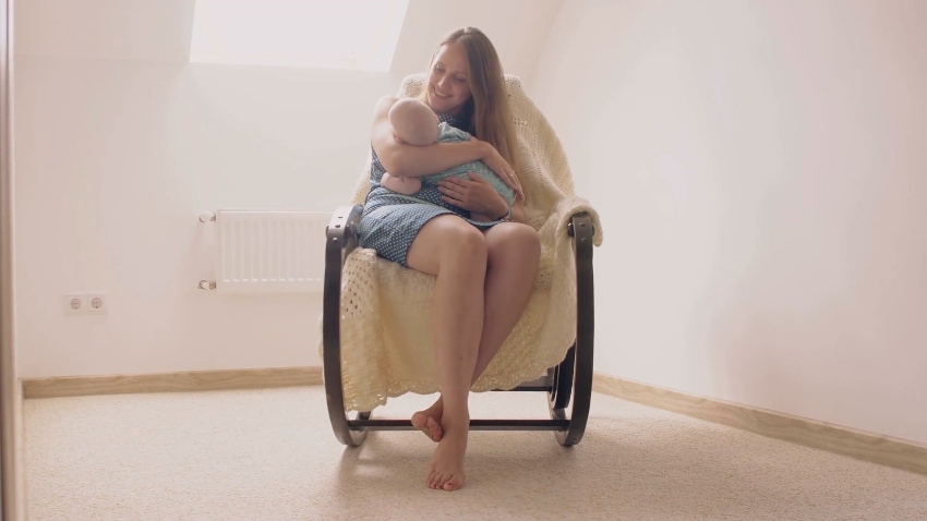 A blonde haired woman sits in a rocking chair gazing down and smiling at a baby in her lap