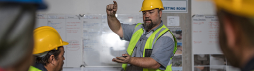 Instructor wearing a yellow hard hat and reflective safety vest gestures while explaining information on a whiteboard to a group of workers in a training setting.