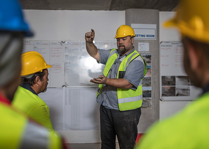Instructor wearing a yellow hard hat and reflective safety vest gestures while explaining information on a whiteboard to a group of workers in a training setting